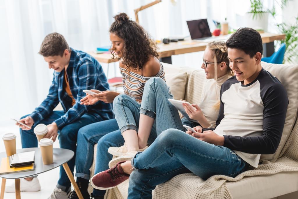 happy multicultural teens sitting on sofa with digital devices